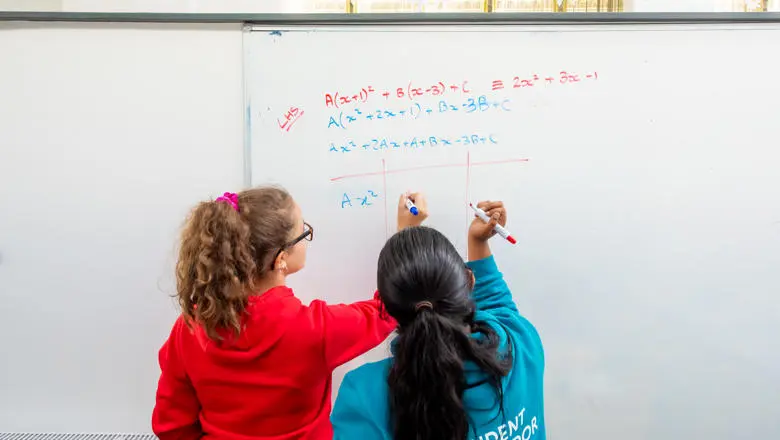 Two young students in casual hoodies collaborating on a complex algebraic equation written on a large whiteboard
