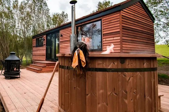 Woman relaxing in a steaming wooden hot tub on a deck next to the Coddiwomple glamping cabin