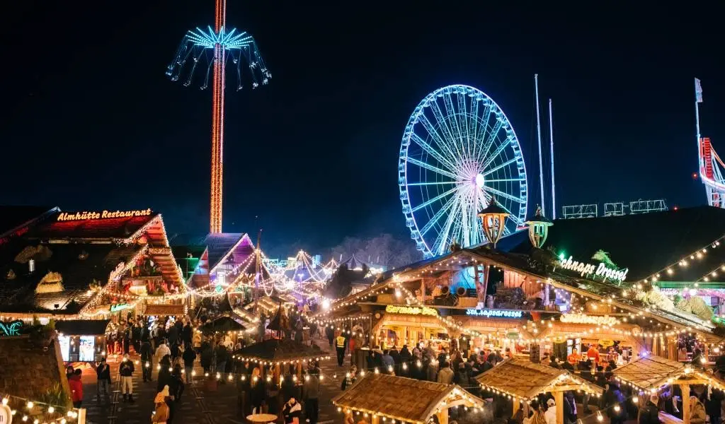 A wide view of Hyde Park Winter Wonderland at night, with glowing market stalls, a Ferris wheel, and a tall swing ride