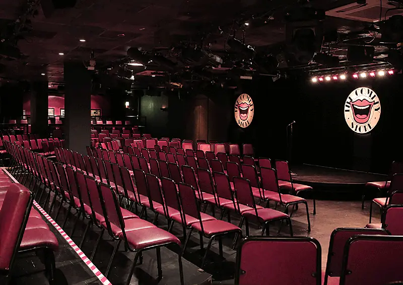 Empty interior of The Comedy Store showing rows of red seats facing the stage with illuminated logos