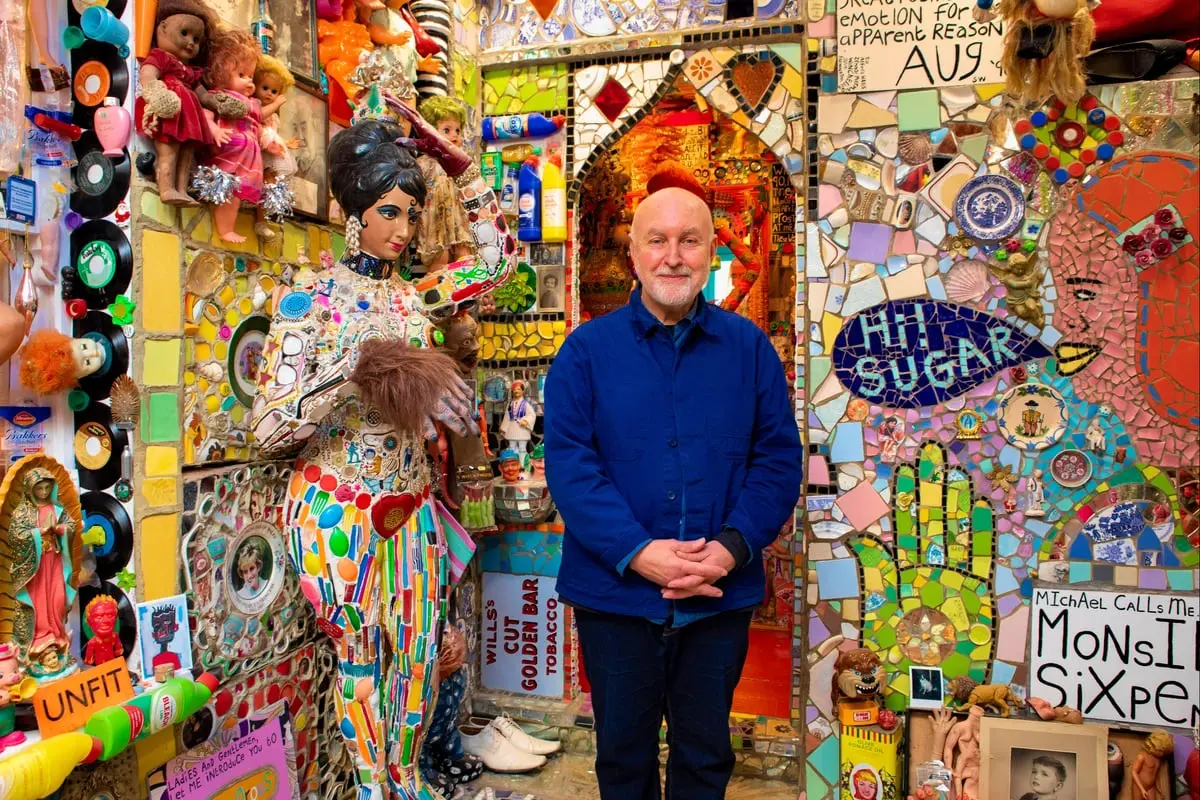 Artist Stephen Wright standing in a room covered with colourful mosaics and found objects at the House of Dreams Museum