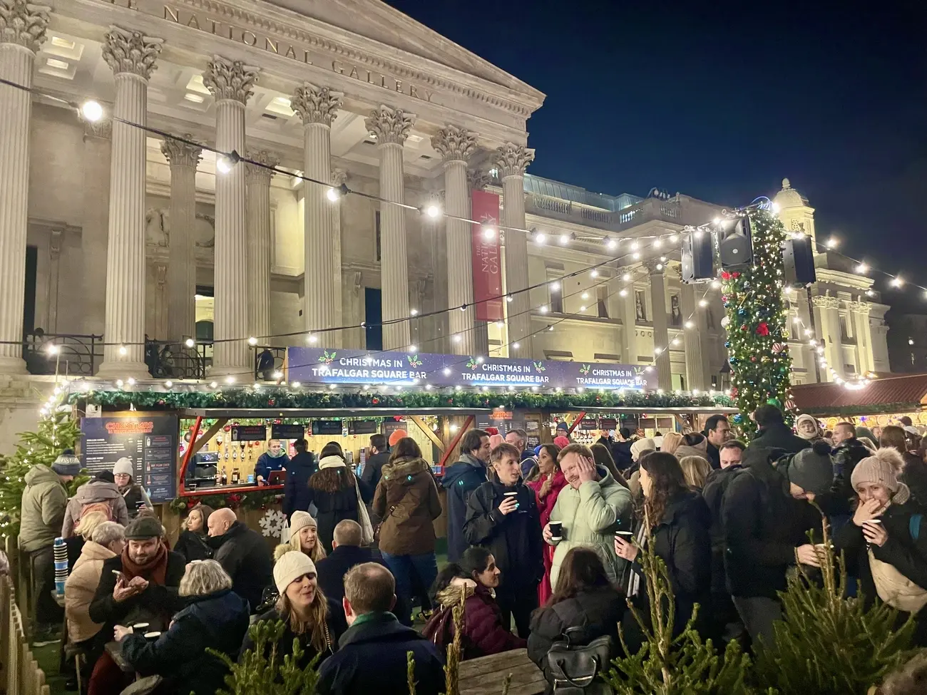 A crowd of people enjoying drinks at the bustling outdoor bar of the Trafalgar Square Christmas Market at night