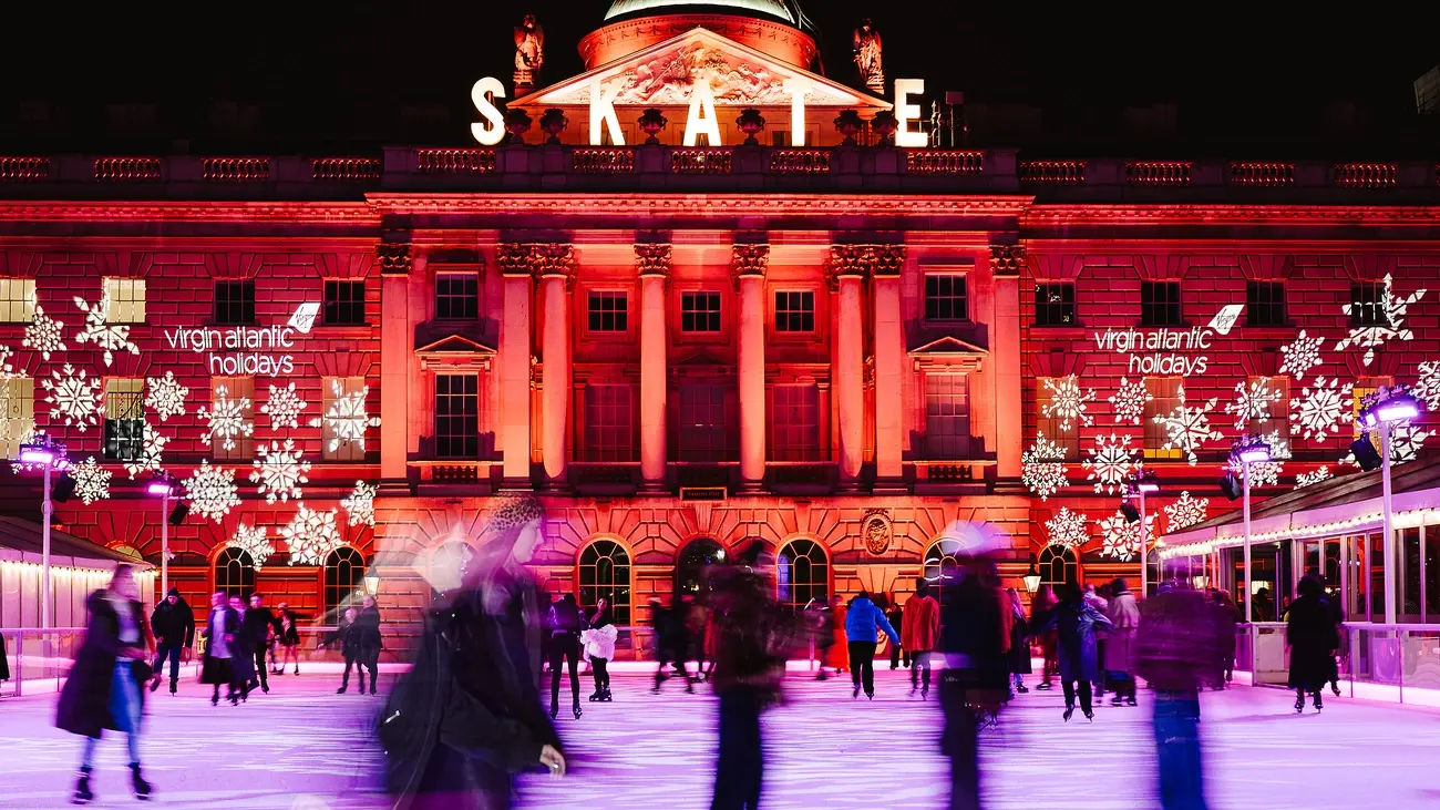People ice skating at night in the grand courtyard of Somerset House, which is illuminated in red with snowflake projections