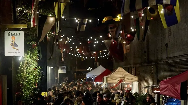 A bustling crowd at the Maltby Street Night Market, which is decorated with string lights and festive flags overhead