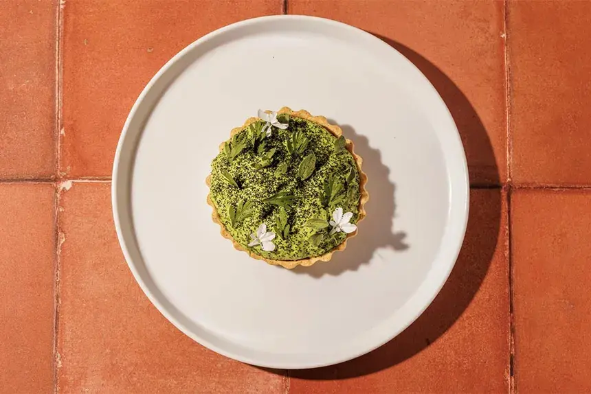 An elegant green vegan dessert tart topped with fresh mint and white edible flowers on a white plate