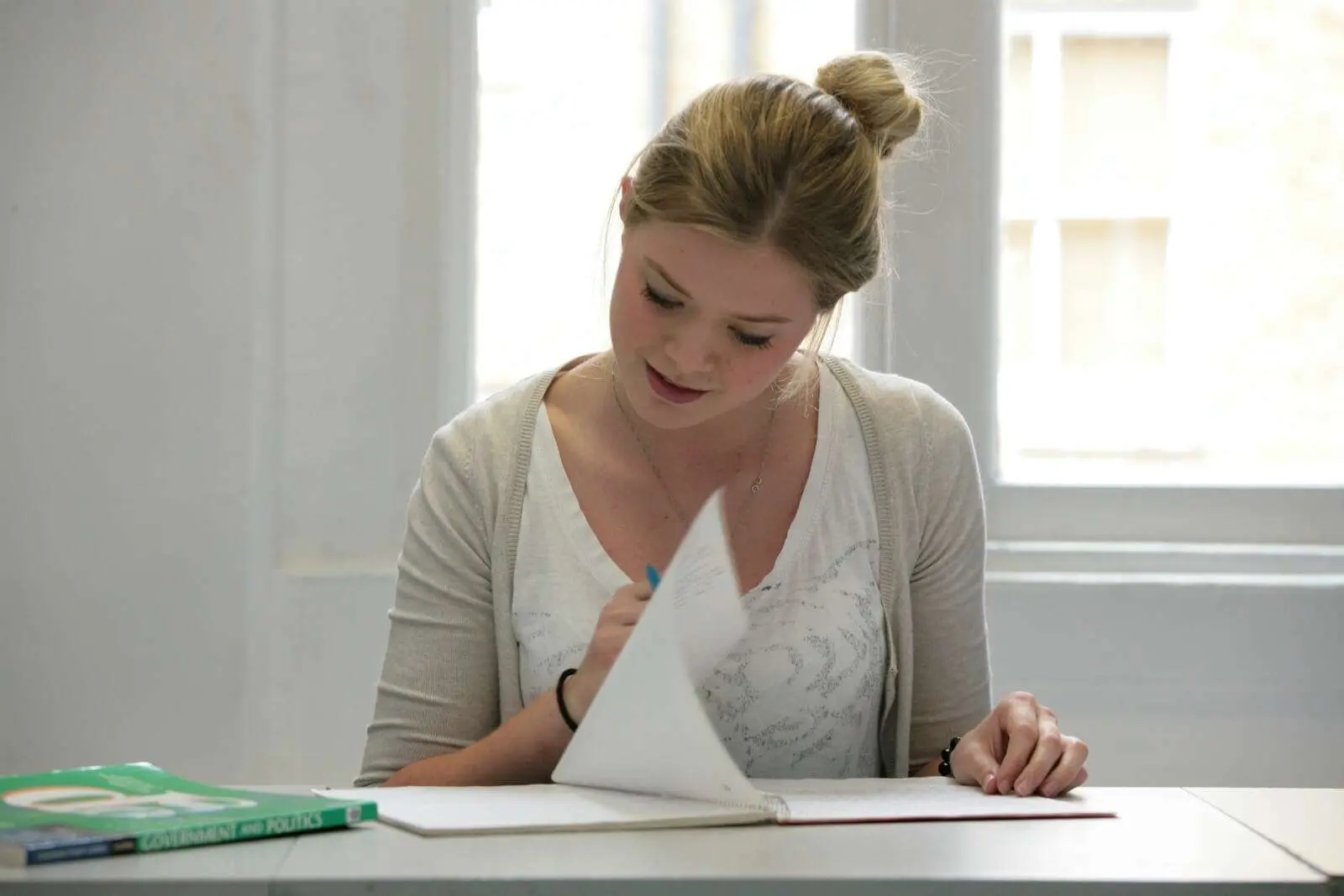 Young female student with blonde hair in a bun, attentively reading and writing in a notebook at a desk