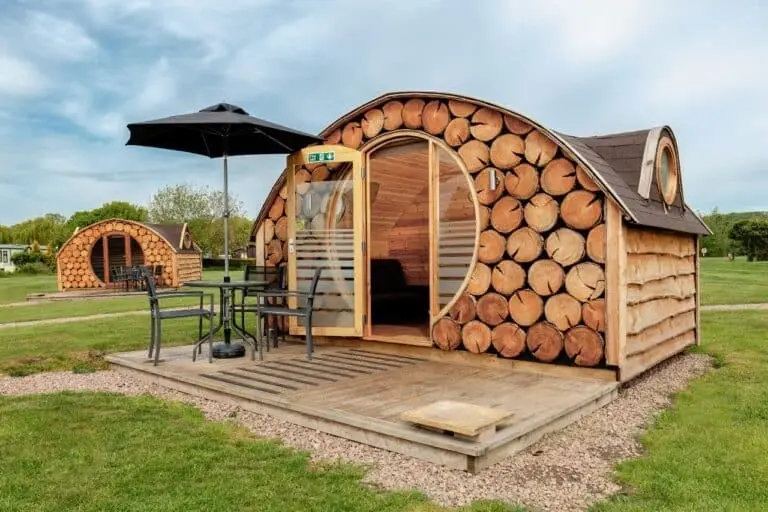 Unique log-effect glamping pod with a circular door, patio table, chairs, and a parasol on a wooden deck