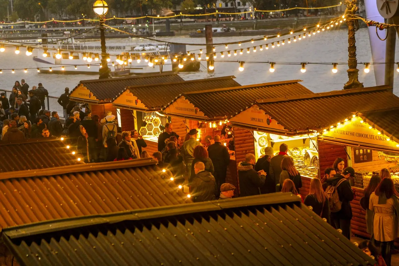 Crowds gather around warmly lit wooden stalls at the Southbank Centre Winter Market, with the River Thames in the background