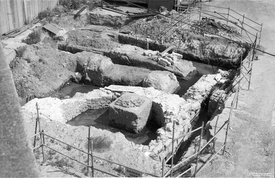 Black and white photo of an archaeological dig site showing exposed stone foundations and trenches, surrounded by scaffolding
