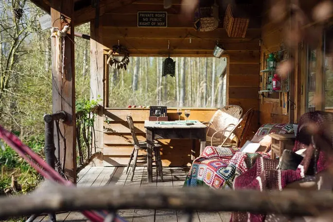 Cosy wooden glamping cabin porch with colourful blankets, wicker chairs, and a view into the woods