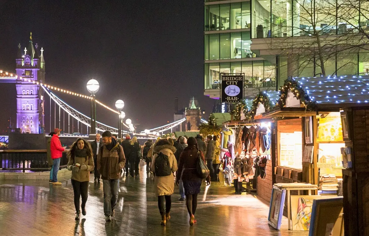 People strolling along the wet pavement at the Winter by the River market, with an illuminated Tower Bridge in the background