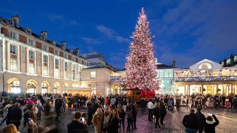A towering, illuminated Christmas tree stands in the crowded piazza of Covent Garden's Christmas market at dusk