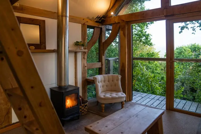 Rustic interior of Uplands Treehouse with a lit wood-burning stove, comfortable chair, and large windows overlooking trees