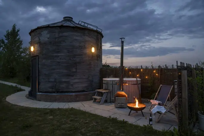 Converted metal grain silo glamping unit at dusk, with an outdoor hot tub, fire pit, and string lights