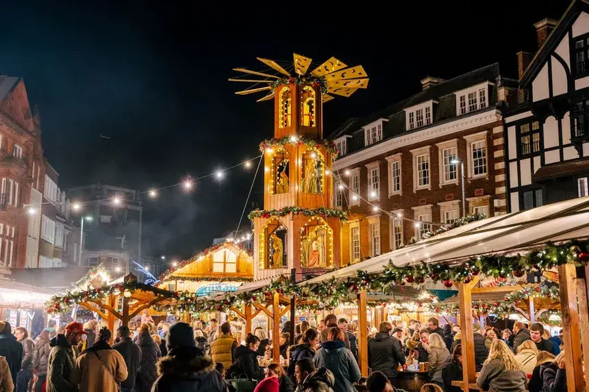 A busy night scene at Kingston Christmas Market, showing crowds around stalls and a traditional German Christmas pyramid lit up