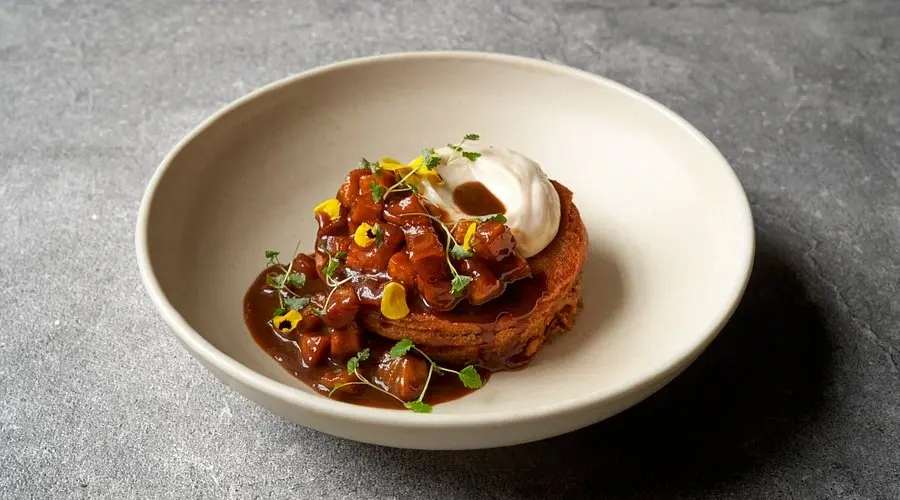 A beautifully plated gourmet vegan dish with a rich sauce and yellow flower garnish in a ceramic bowl