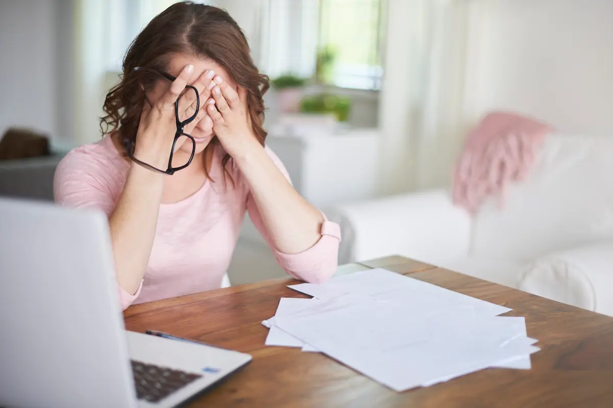 Woman looking stressed with head in hands sits at a table with laptop and papers