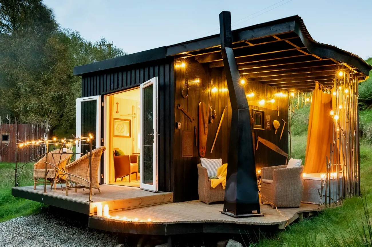 Modern black Arcadia glamping cabin with a wooden deck, wicker chairs, and warm string lights in the evening