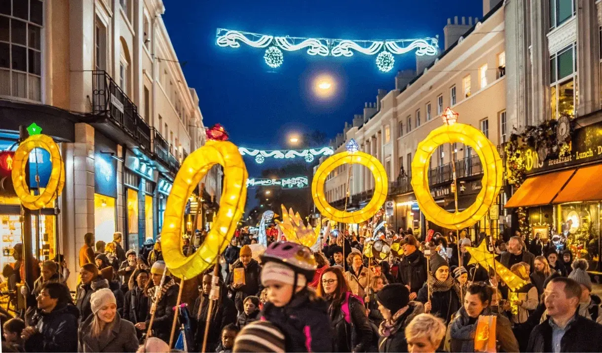 A crowd of people, including children, participating in the Greenwich Christmas Lantern Parade under festive street lights