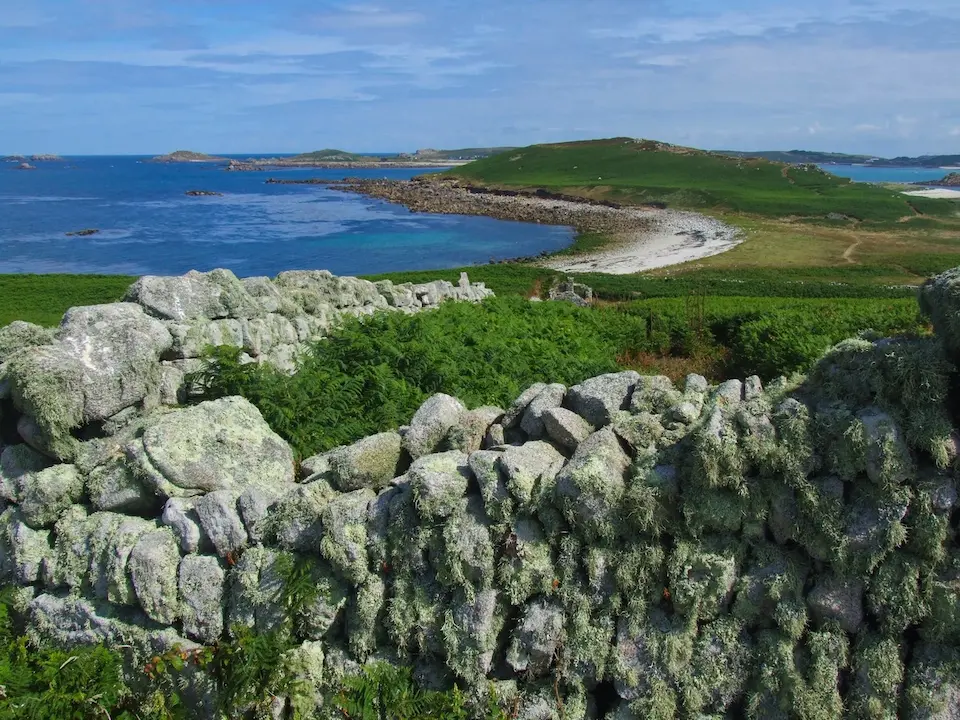 View over a lichen-covered ruined stone wall towards the green hills and blue sea of Samson Island, Isles of Scilly