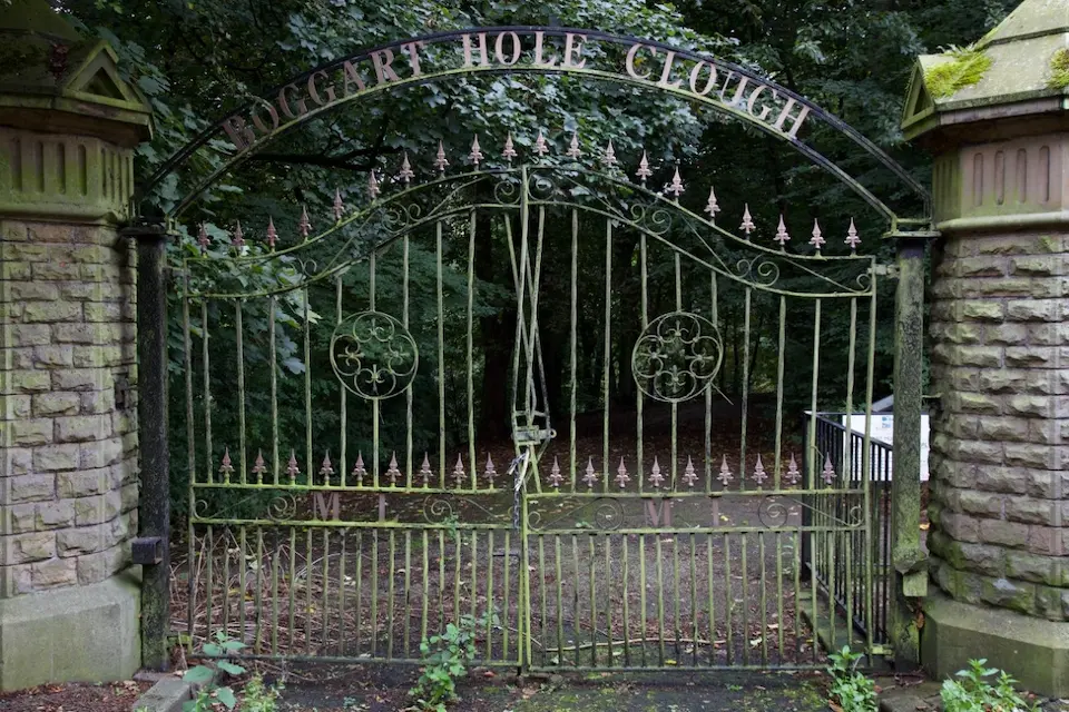 Ornate, weathered green iron gate with 'Boggart Hole Clough' written above, flanked by stone pillars, leading into woods